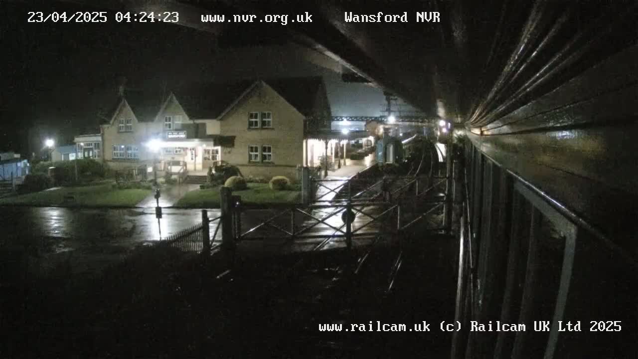 A nighttime view of a wet train station with a two-story building and a crossing gate.
