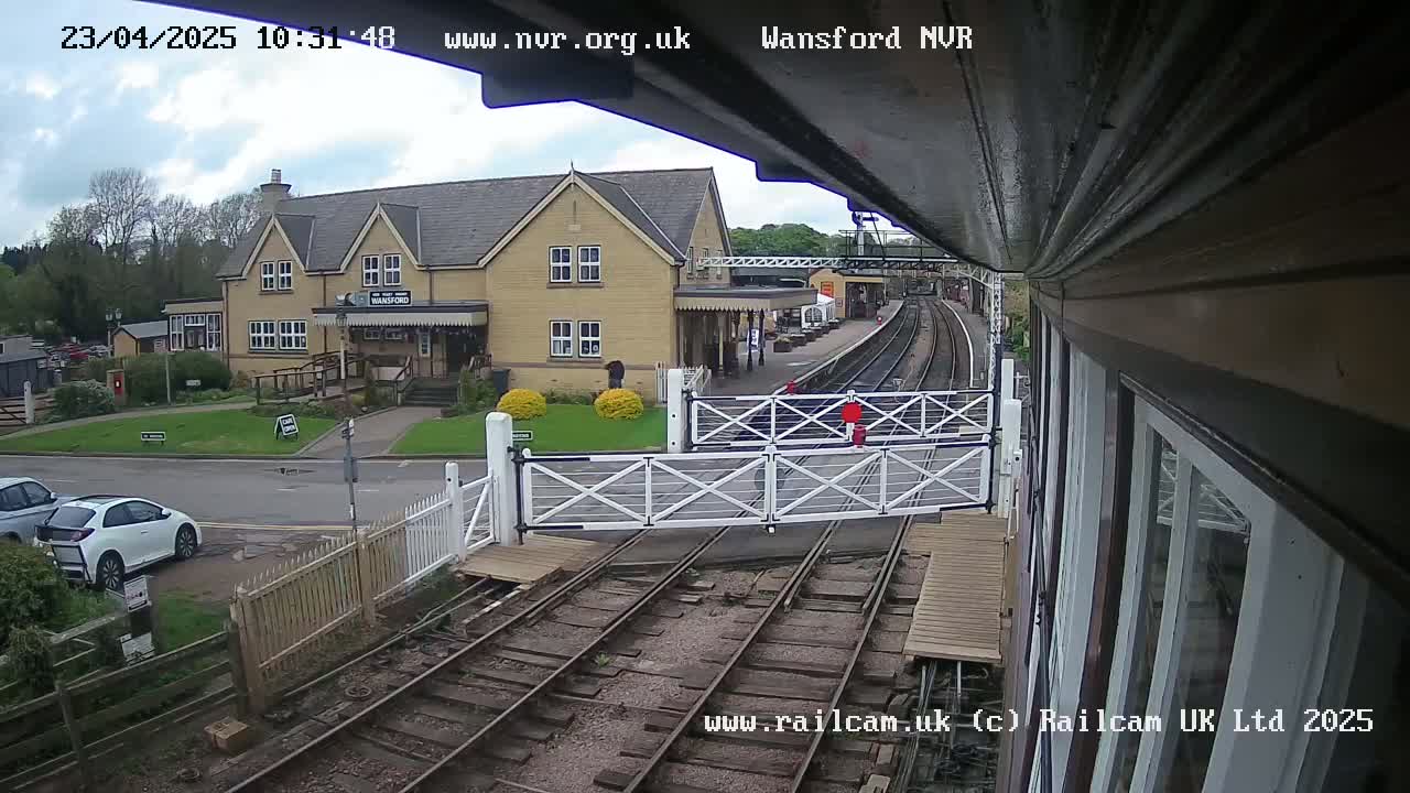A partly cloudy day at a train station shows a two-story building, railway tracks with crossing gates down, and a few cars parked nearby.