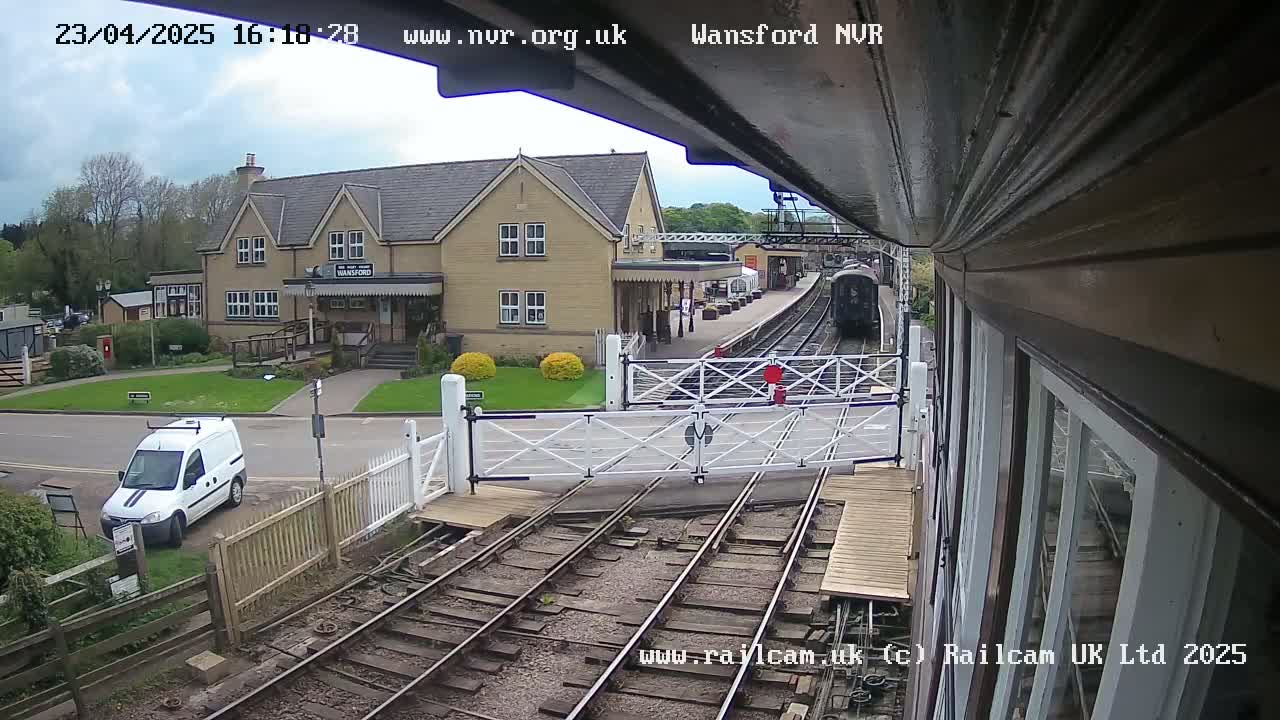 A train approaches a station on a partly cloudy day, passing through a level crossing with a white van parked nearby.