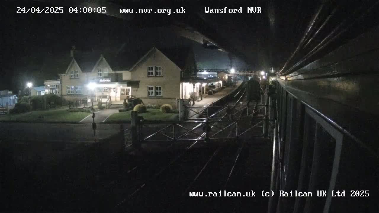A nighttime view from a train shows a two-story building and train station platform with tracks leading away.