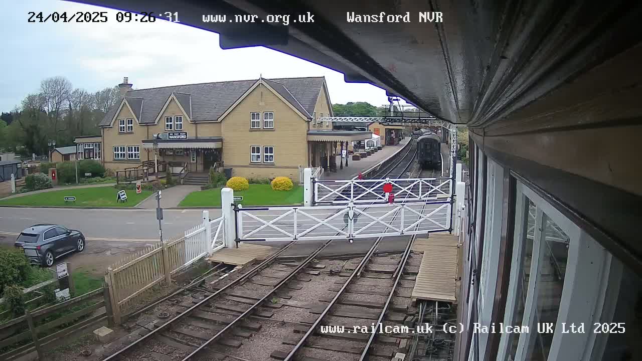 A train is visible on the tracks near a railway station building and a car is parked on the road outside.