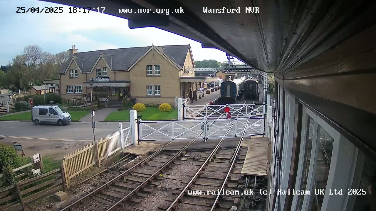 A train is stopped at a railway station on a partly cloudy day, with a van parked nearby and a two-story building visible in the background.