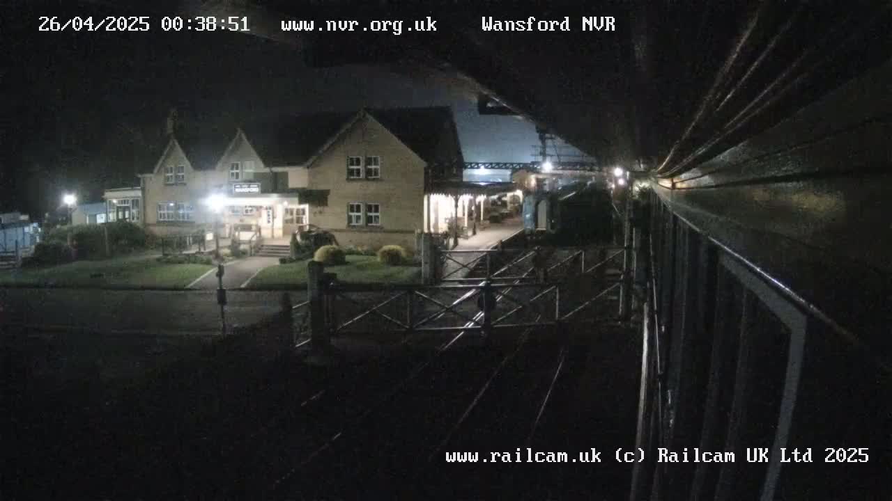 A nighttime view from a train shows a two-story building with lights on, a train station platform, and a crossing gate.
