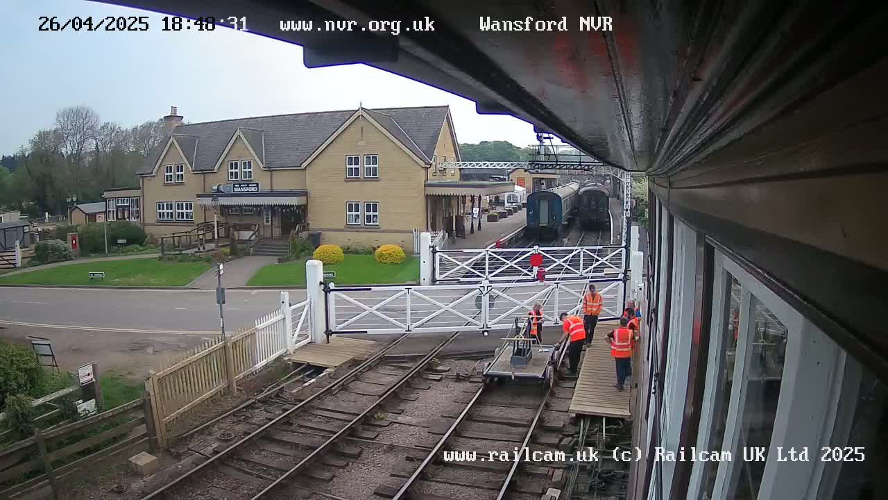 A train station with a two-story building, a railway crossing with a few workers, and a train parked on the tracks under a partly cloudy sky.