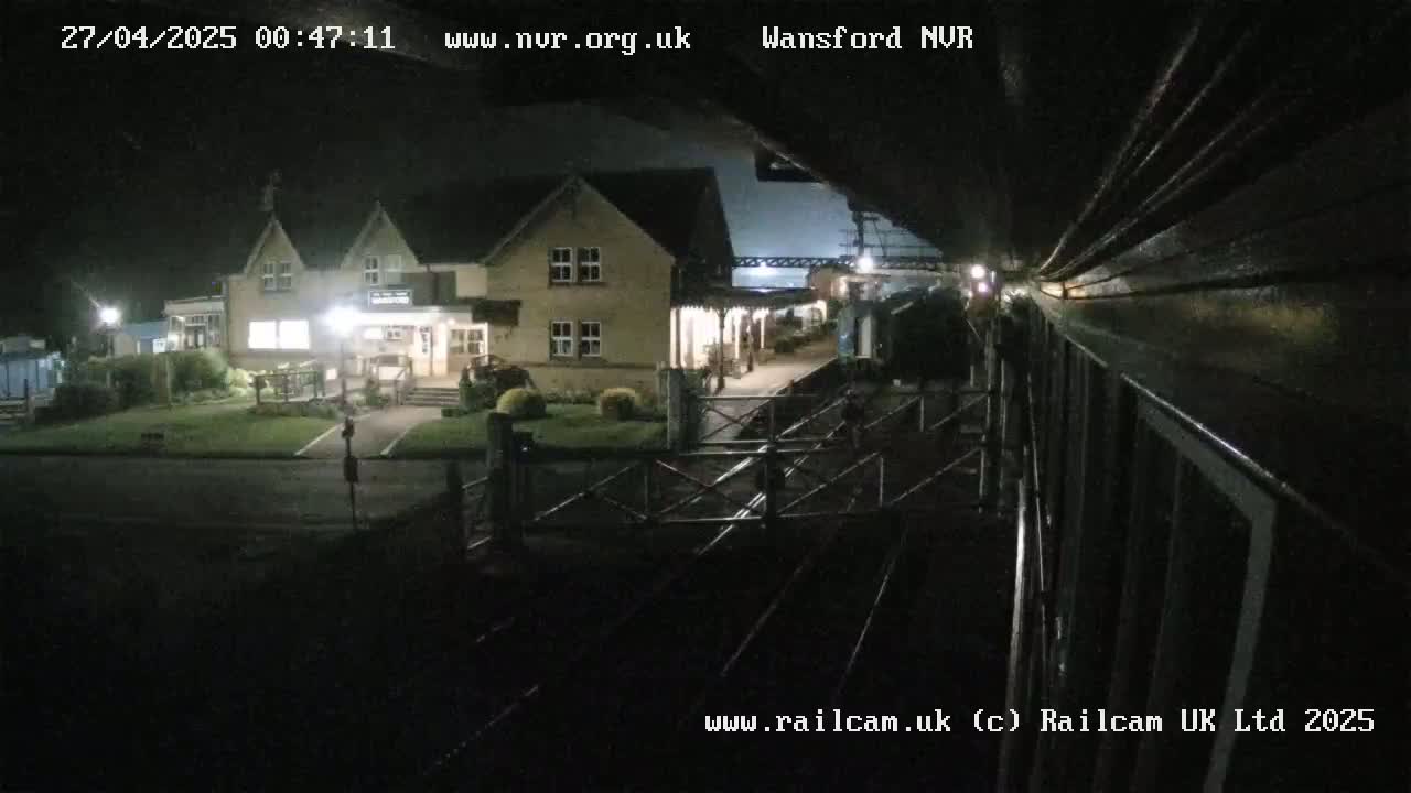 A nighttime view from a train shows a two-story building with lights on near a train station, with train tracks in the foreground.