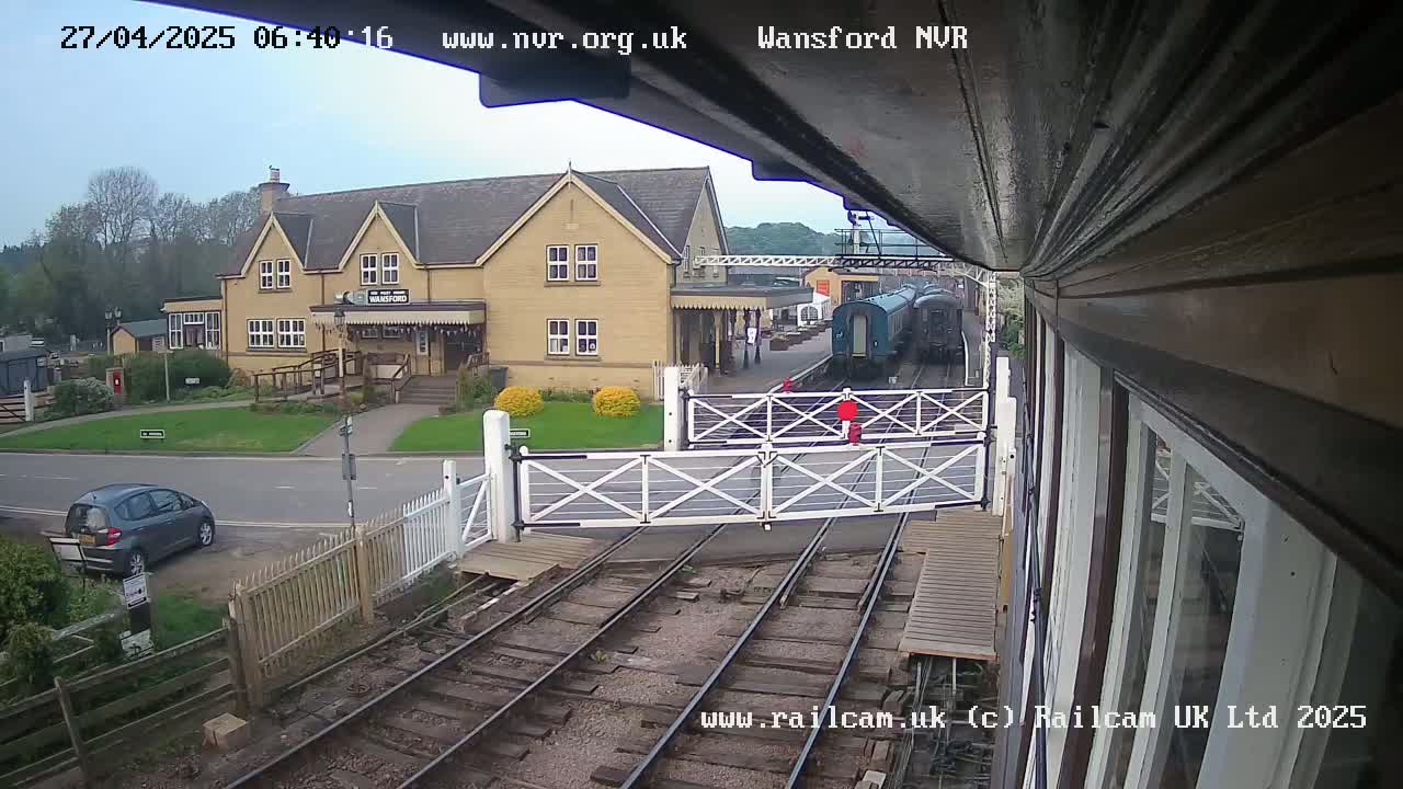 A train is stopped at a station on an overcast day, with a car parked nearby and a two-story building visible in the background.