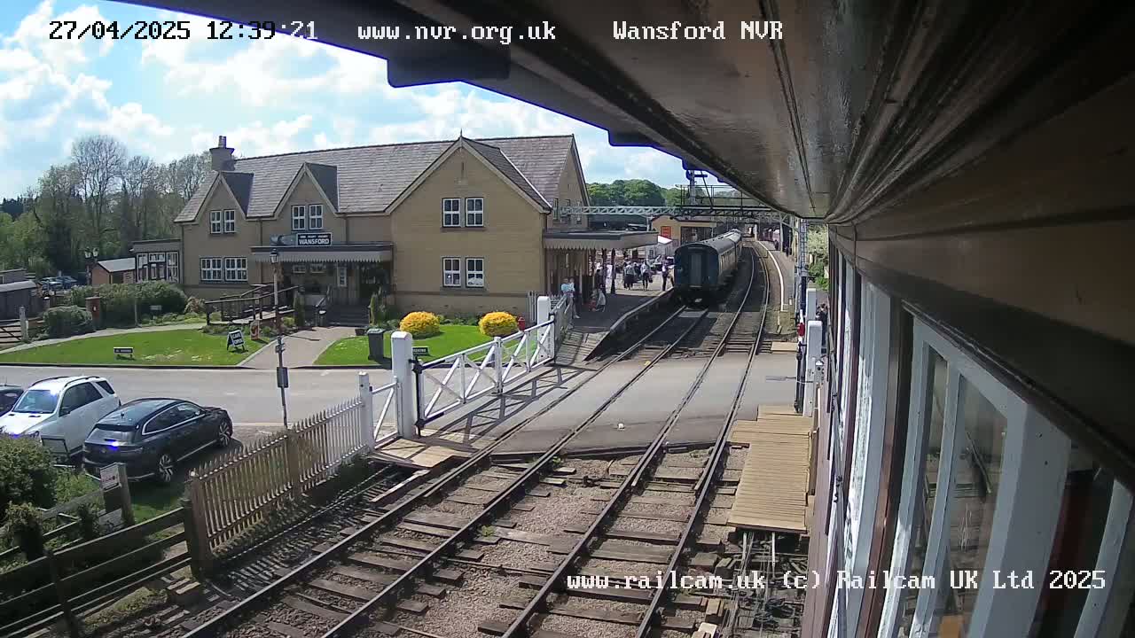 A train pulls into a station on a sunny day, with a two-story building and several cars parked nearby.