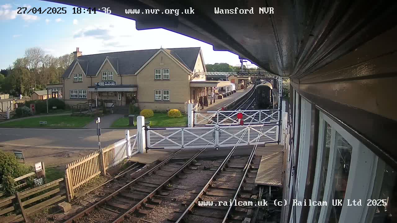A train approaches a railway station under a partly cloudy sky, with a building and crossing gates visible in the foreground.