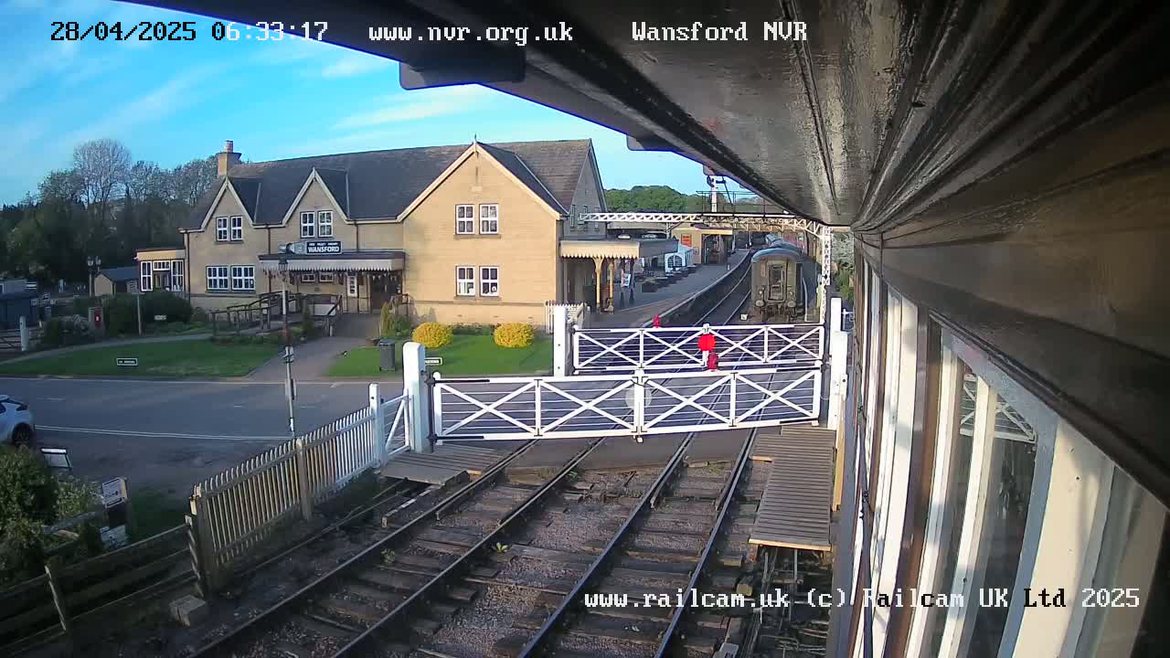 A train approaches a station under a partly sunny sky, passing through a level crossing with white gates.