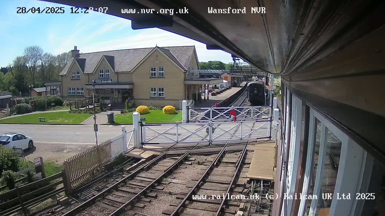 A sunny day at a train station shows a steam train arriving near a station building, with a white car parked nearby and a level crossing gate in the foreground.