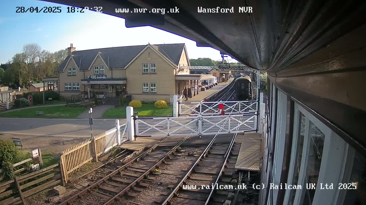 A train is arriving at a railway station on a sunny day, with a building and crossing gates visible.