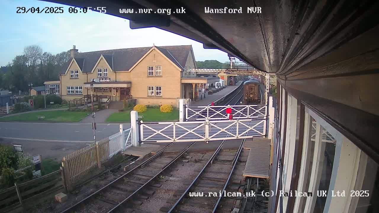A train is arriving at a railway station on a partly cloudy day, passing through a level crossing in front of a building.