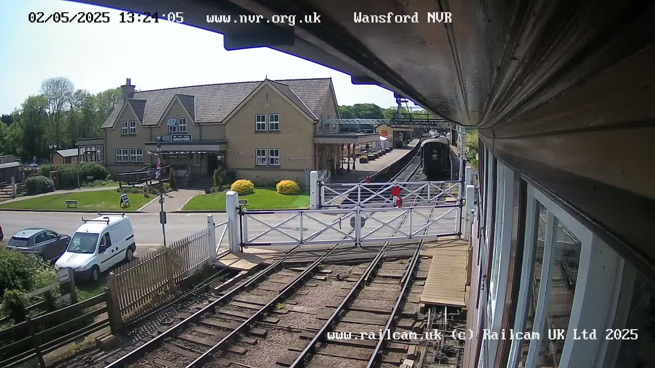 A train approaches a station on a sunny day, passing through a level crossing near a building and several parked cars.