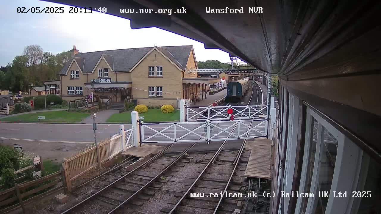 A train is arriving at a station building, with level crossing gates closing, under a partly cloudy sky.