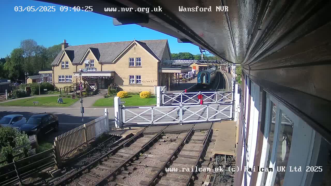 A train is arriving at a station on a sunny day, passing through a level crossing in front of a light-colored building.