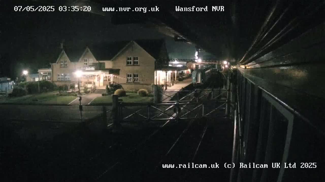 A nighttime view from a train shows a building and train station platform with a train partially visible in the distance.