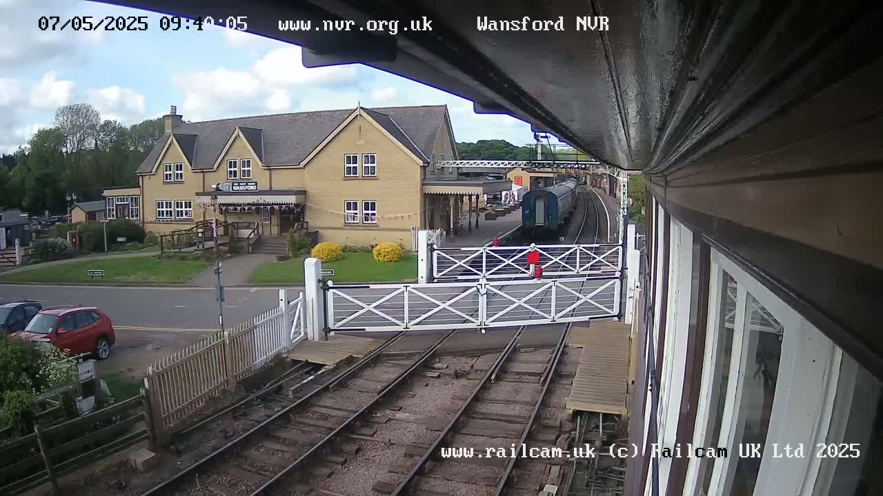 A partly cloudy day at a train station shows a train arriving at a platform near a yellow building, with cars parked nearby and level crossing gates down.