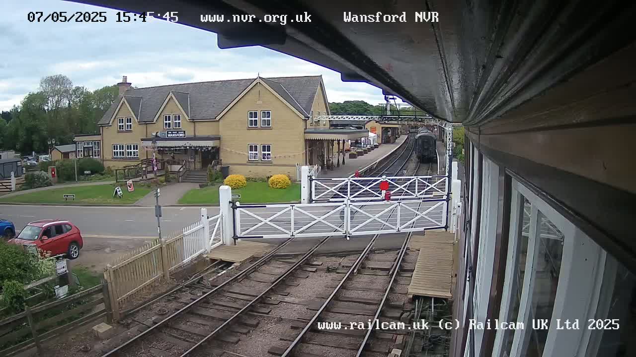 A train approaches a railway station on an overcast day, passing through a level crossing with a nearby building and cars parked on the road.
