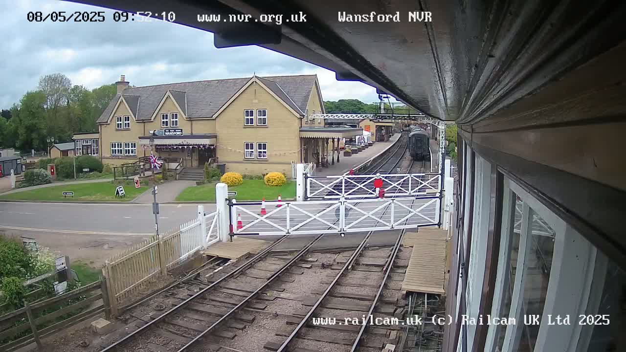 A train approaches a station building on an overcast day, passing through a level crossing with its gates down.
