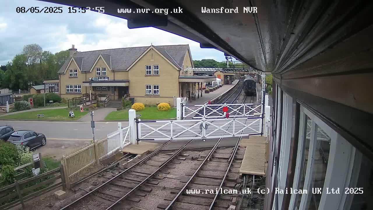 A train passes a station building on a partly cloudy day, with cars parked nearby and level crossing gates down.