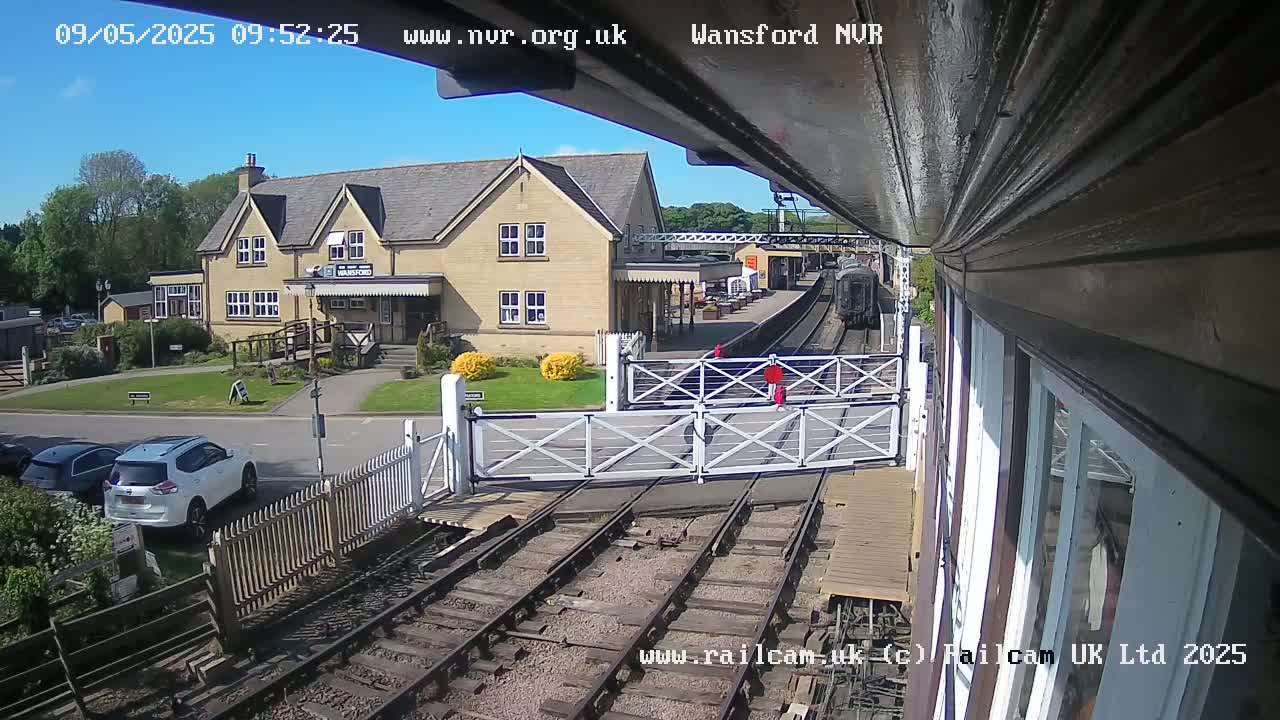 A train approaches a station on a sunny day, passing through a level crossing with cars parked nearby and a two-story building visible in the background.