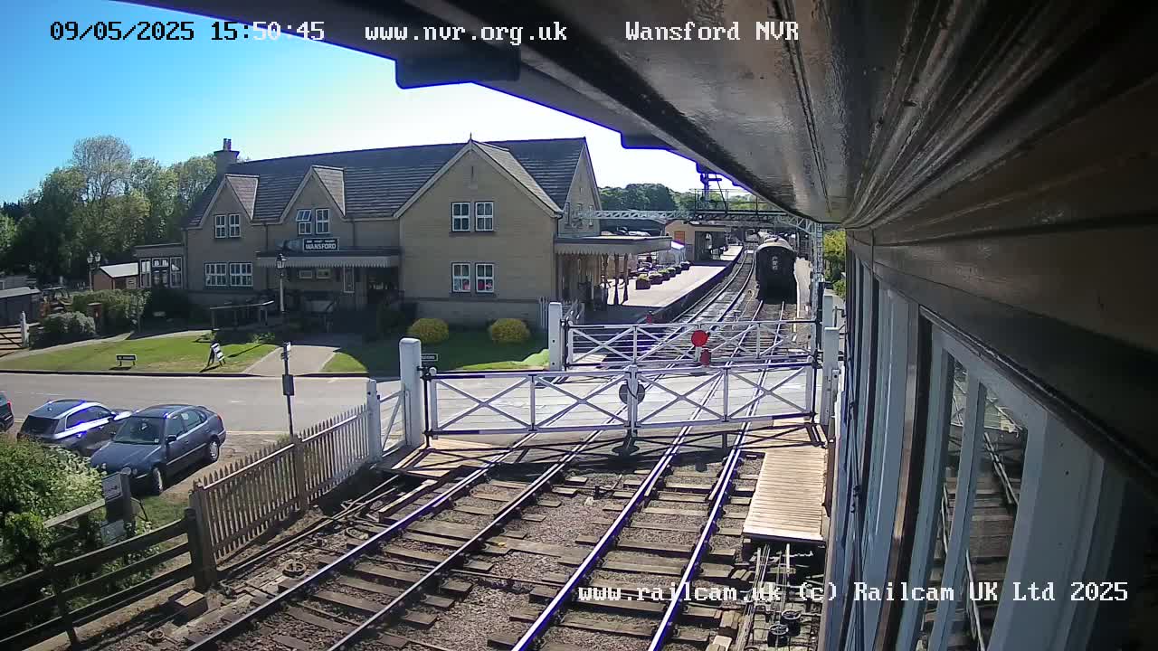 A train approaches a railway station on a sunny day, passing through a level crossing with cars parked nearby and a building visible in the background.
