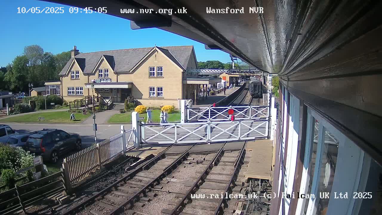 A train is visible at a station on a sunny day, with several cars parked nearby and pedestrians crossing the tracks at a level crossing.