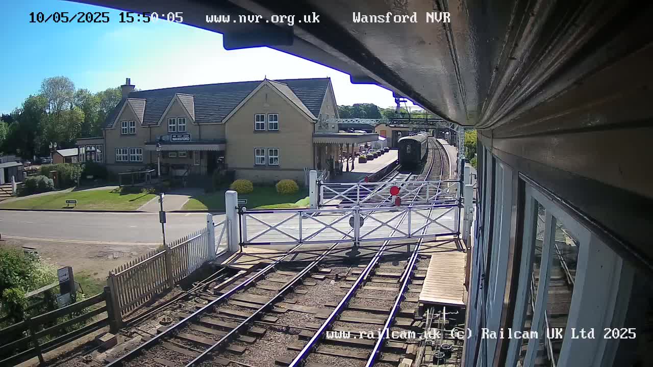 A train approaches a sunny railway station with a two-story building beside the tracks.