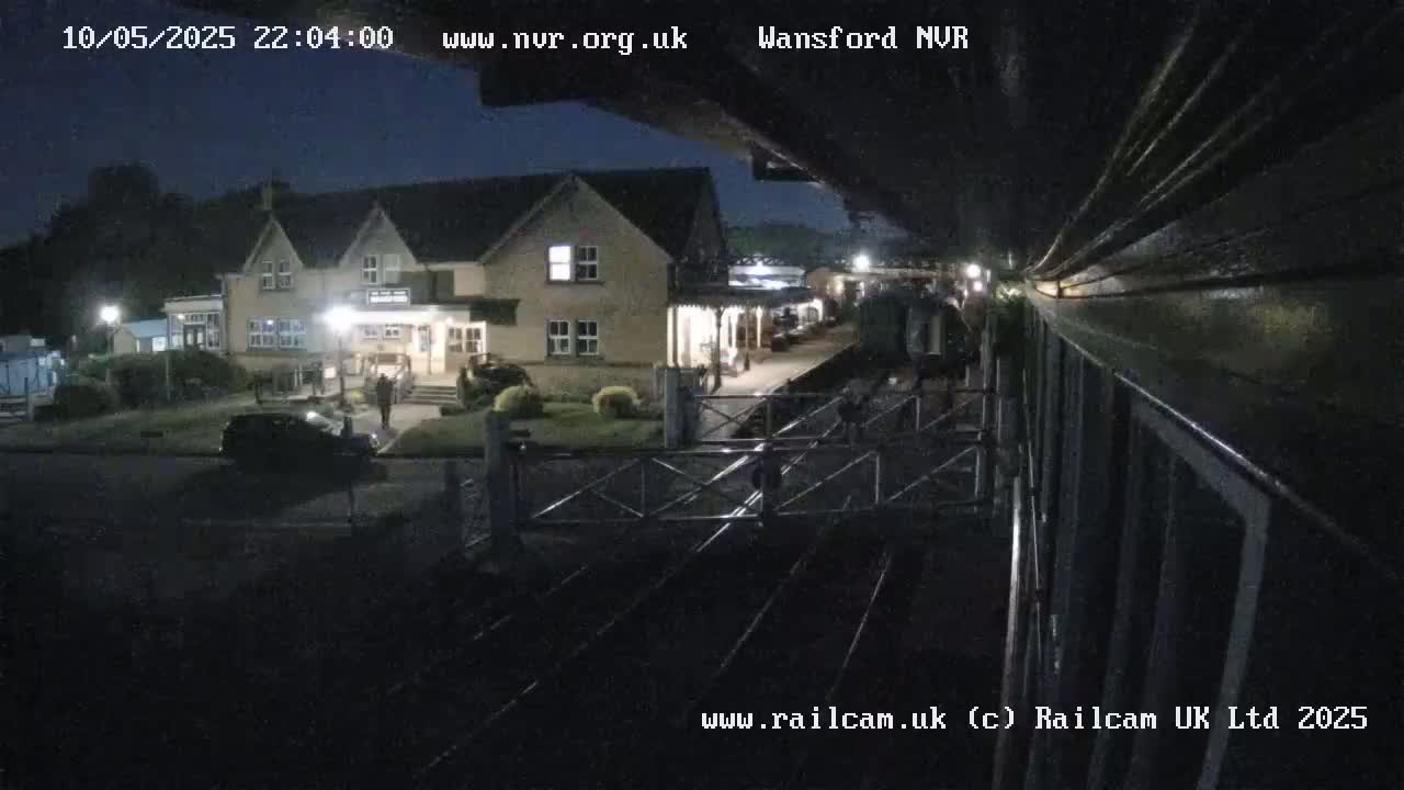 A nighttime view of a train station building and a railway crossing, with cars parked nearby under a dark sky.