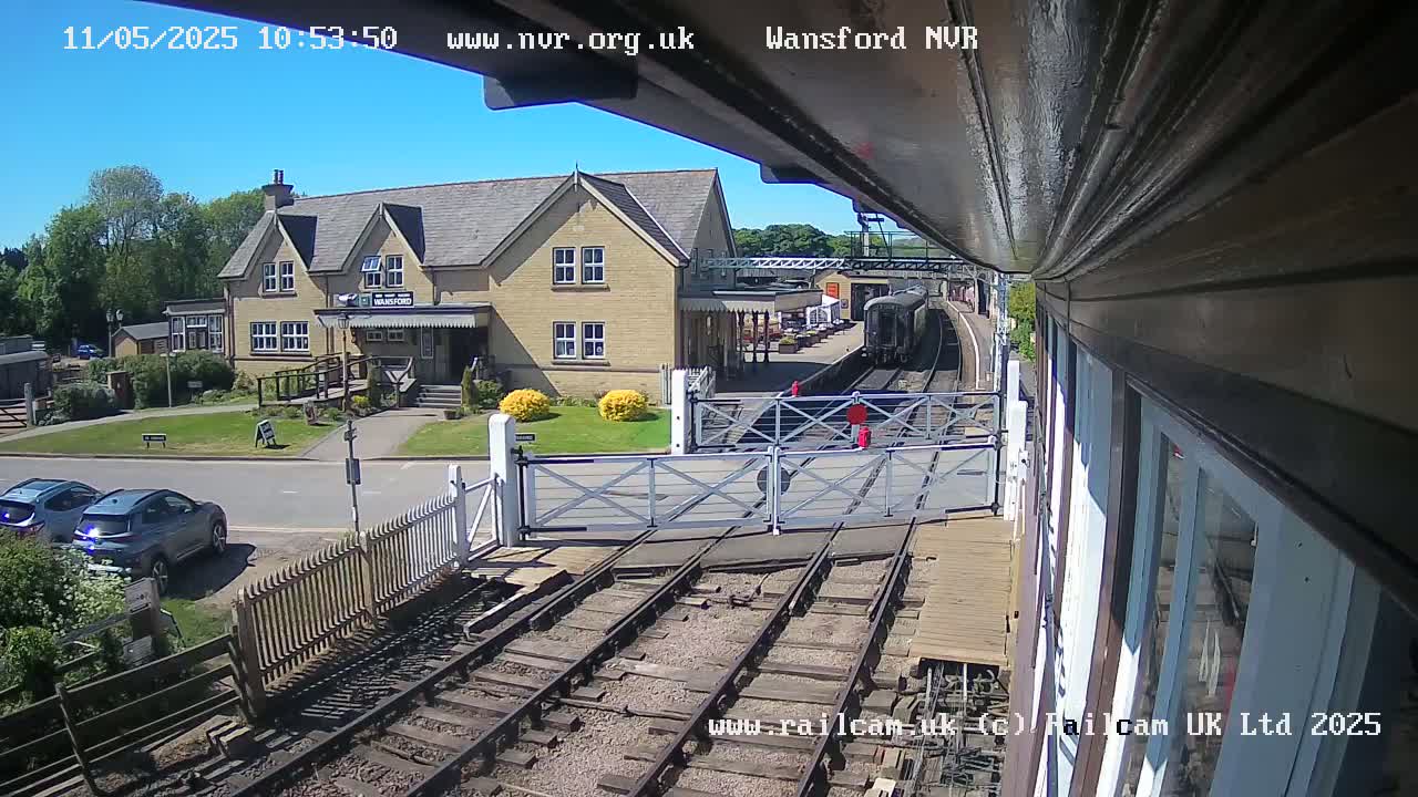 A train is arriving at a station on a sunny day, passing through open crossing gates near a two-story building.