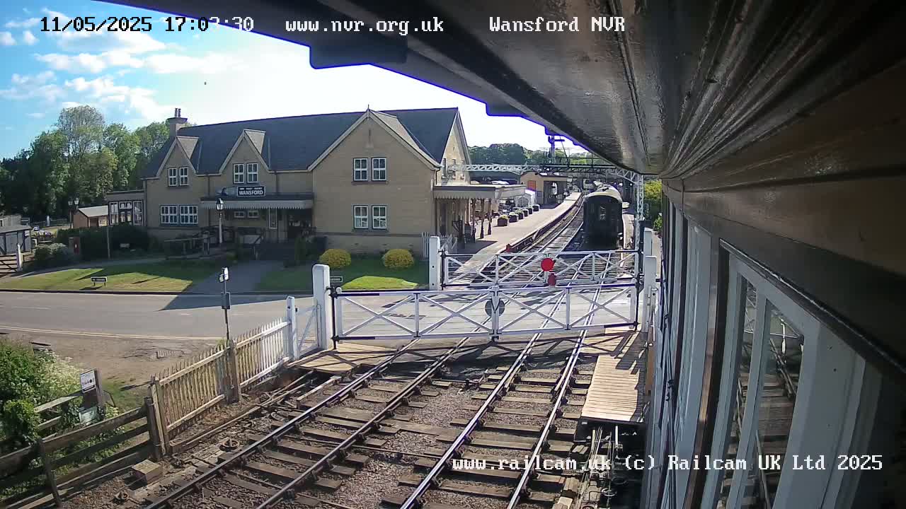 A train approaches a railway station on a sunny day, with a building and level crossing visible.