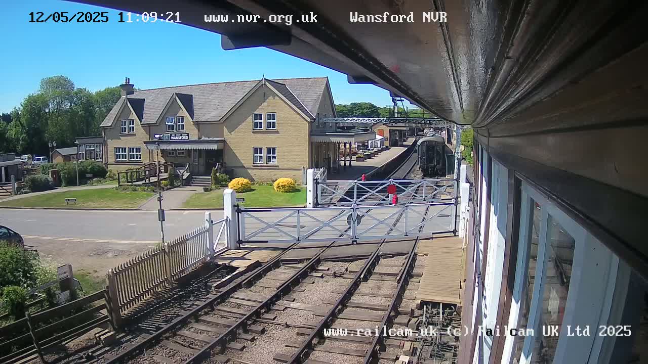 A train approaches a station building on a sunny day, with level crossing gates down.