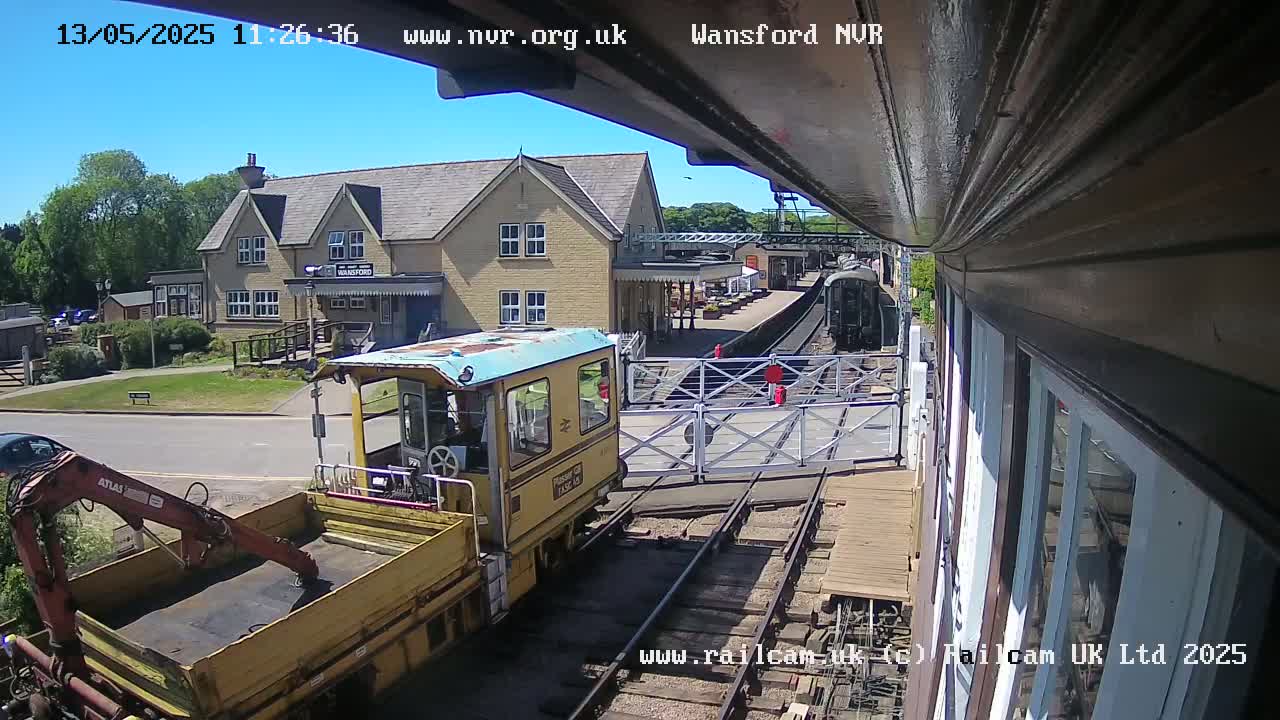 A yellow railway maintenance vehicle is positioned near a railway crossing in front of a station building on a sunny day.