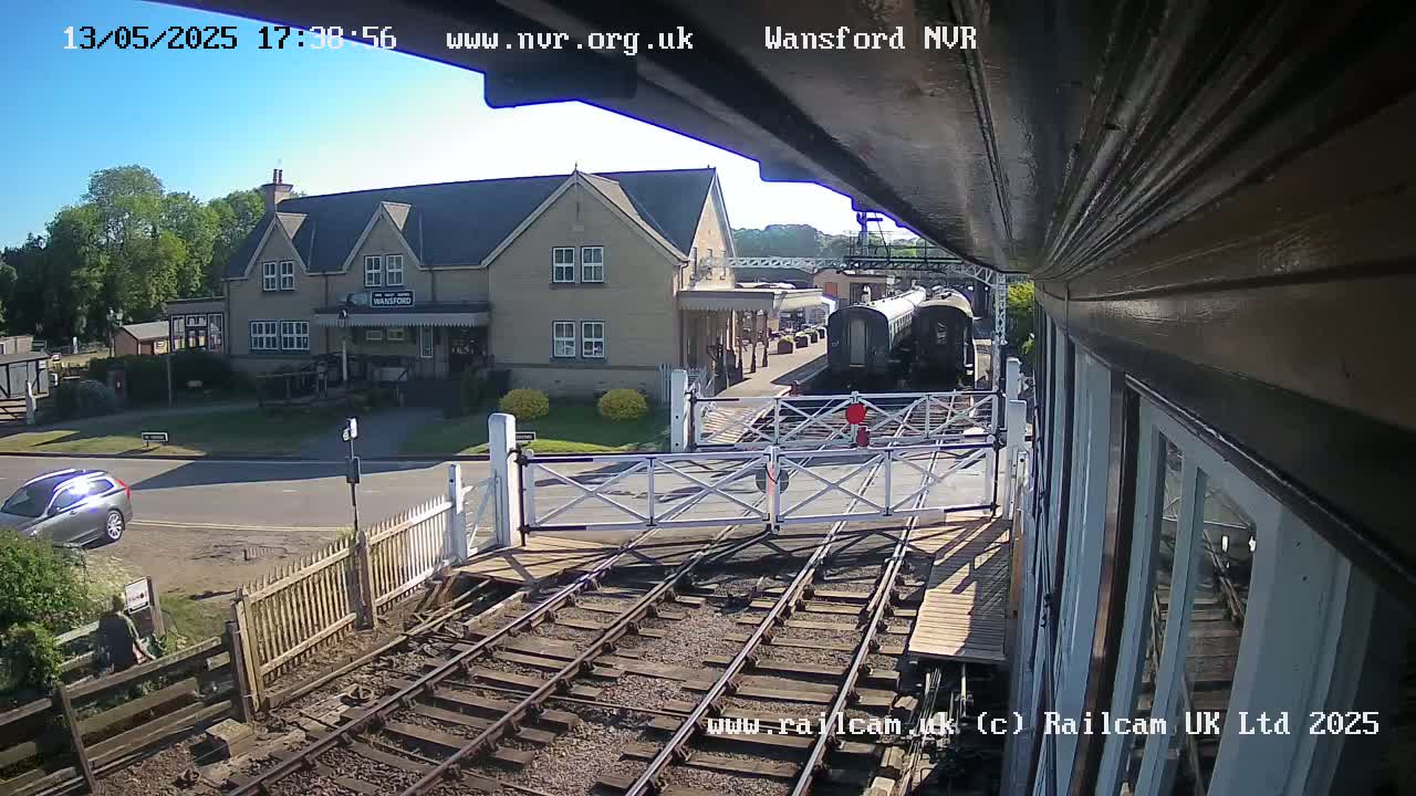 A sunny view from a train platform shows railway tracks, a crossing gate, a station building, and a train.