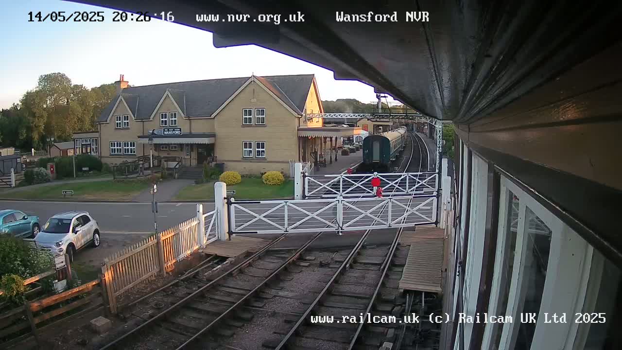 A train is stopped at a station on a clear evening, with two cars parked nearby.