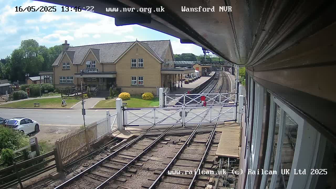 A sunny day view from a train shows a railway station with a two-story building, a level crossing gate, and train tracks.