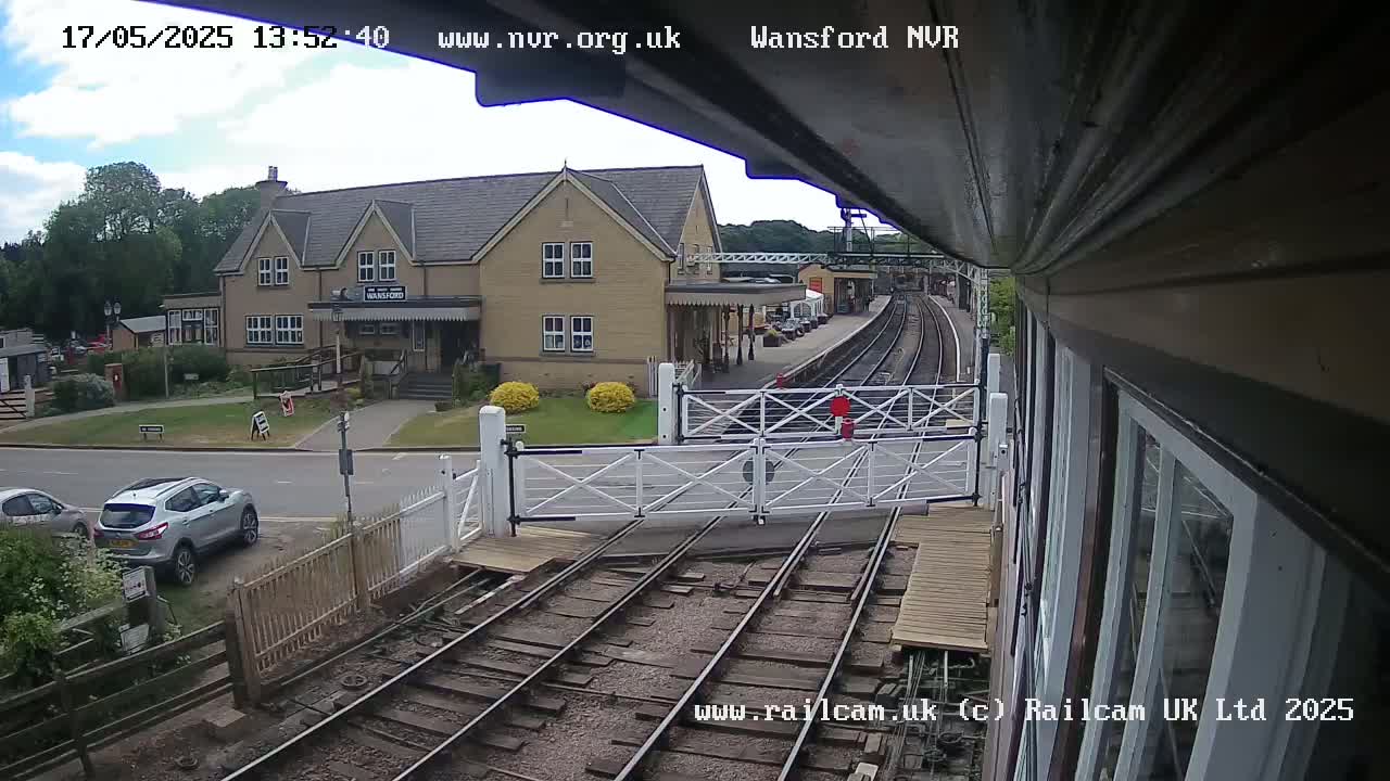 A partly cloudy day shows a train station with a two-story building and railway tracks, alongside a road with parked cars.