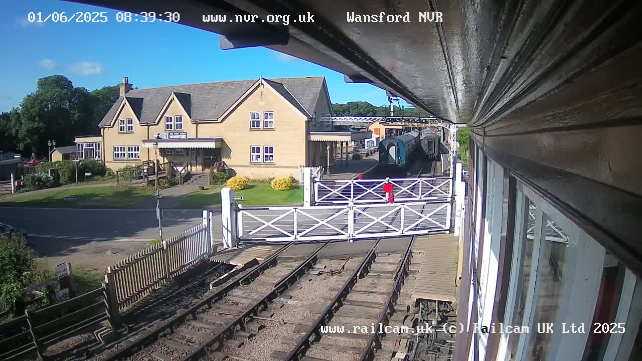A train sits at a station on a sunny day, with a two-story building visible beyond a level crossing.