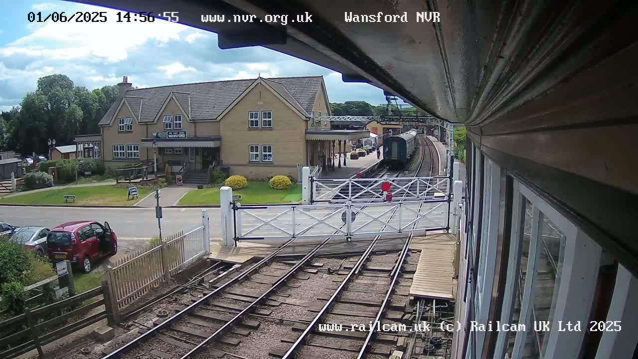 A partly cloudy day at a train station shows a train arriving at a platform next to a building, with a red car parked nearby and level crossing gates down.