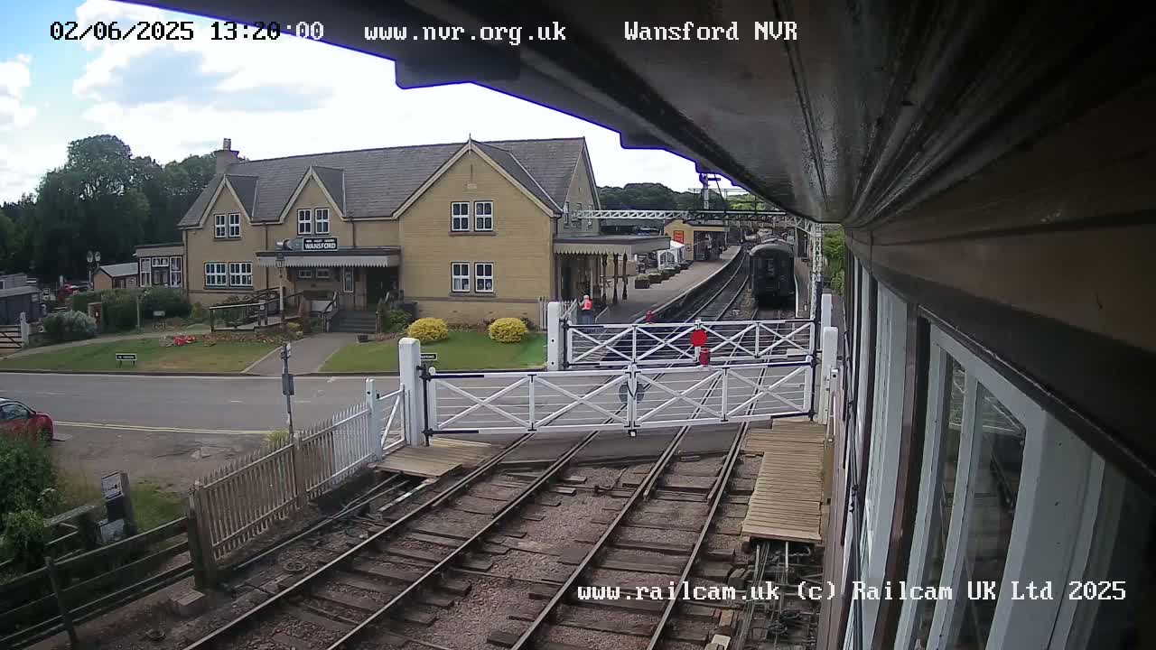 A train approaches a station on a partly cloudy day, passing through a level crossing gate near a two-story building.