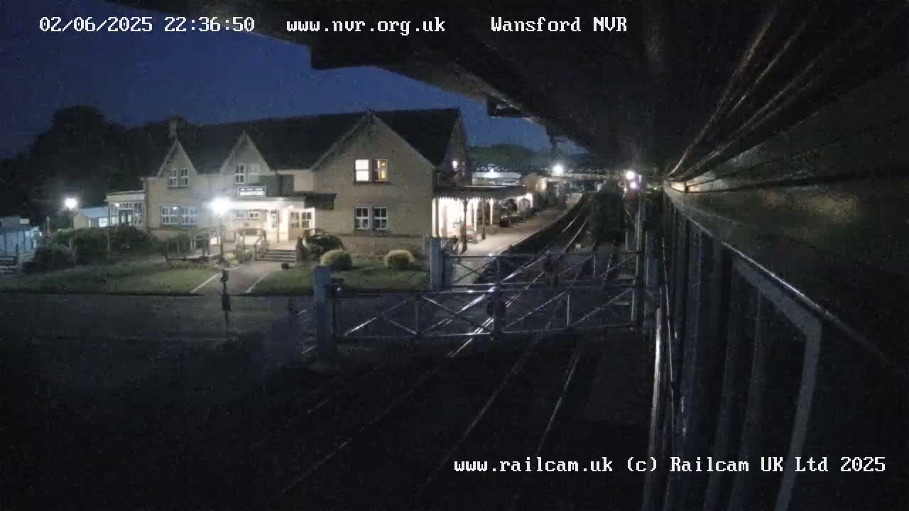 A nighttime view from a train shows a railway station building and tracks under a mostly clear, dark sky.