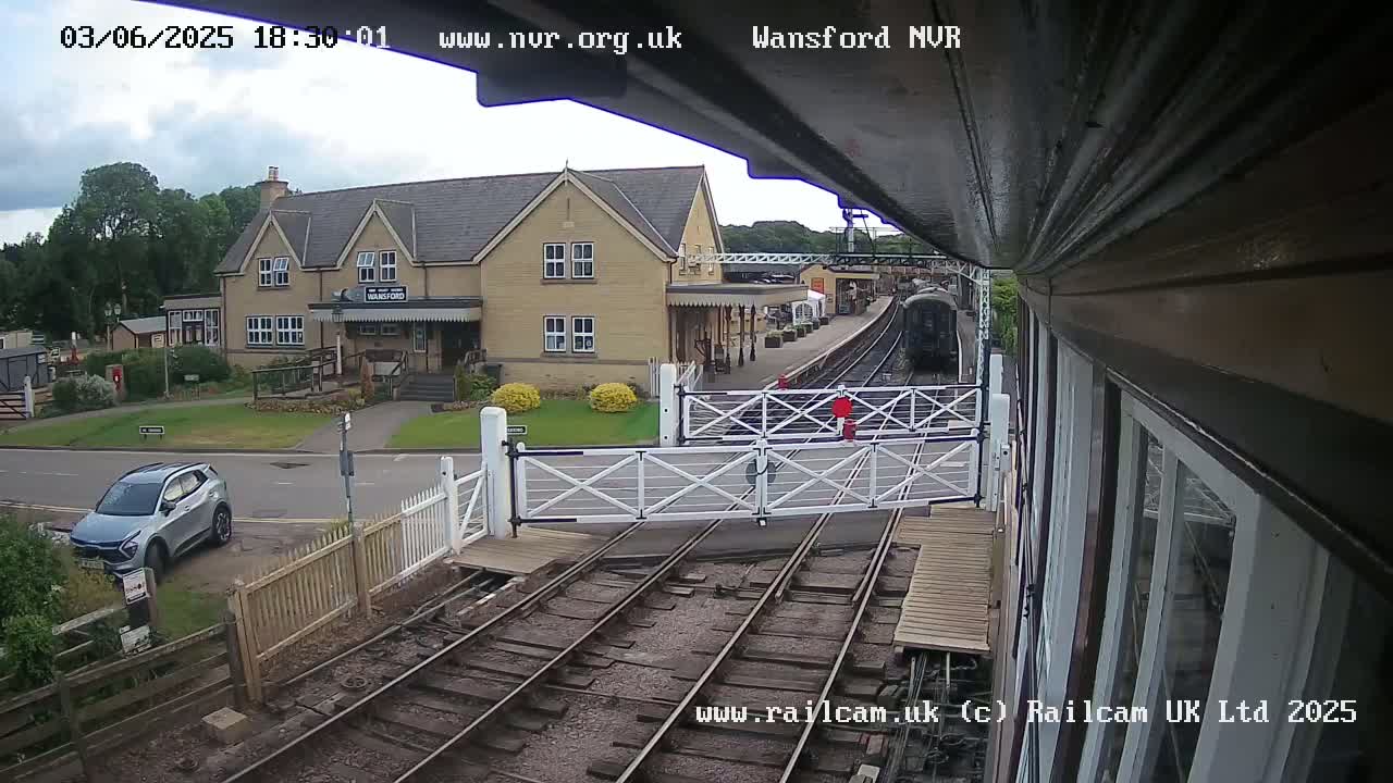 A partly cloudy day shows a train approaching a station with a small gray car parked nearby, and level crossing gates are down.
