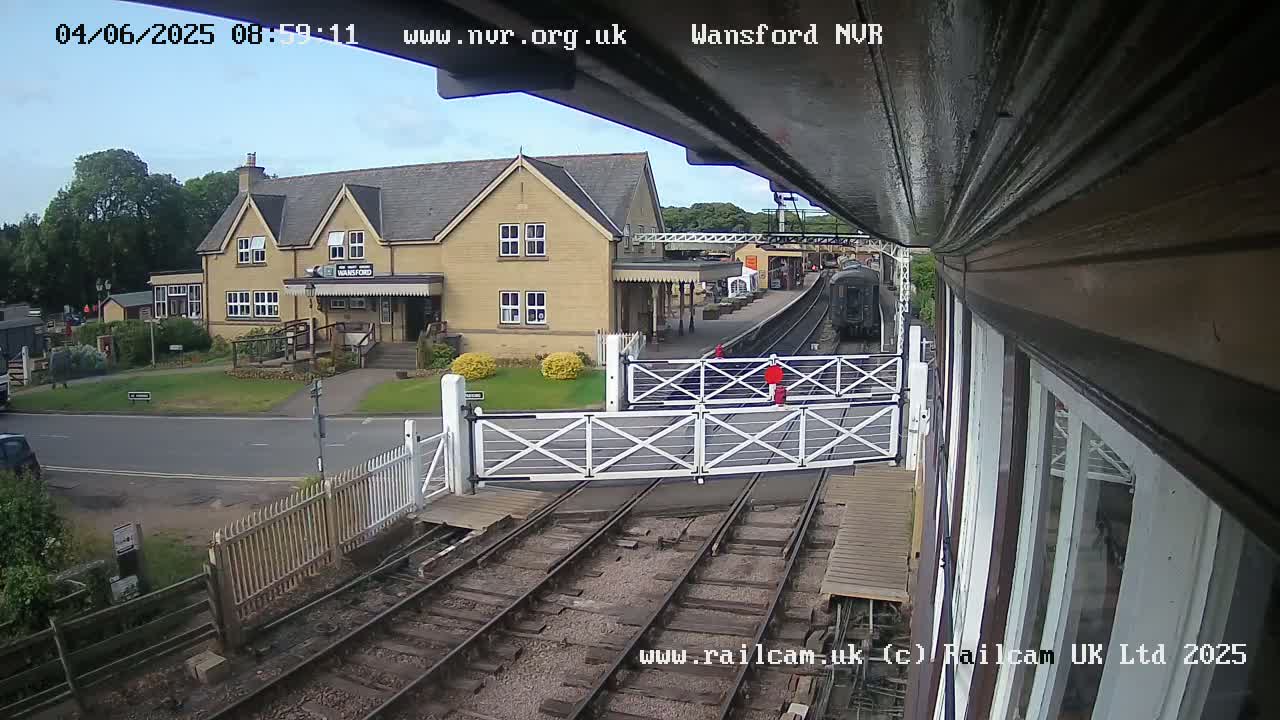 A train is arriving at a station on a partly cloudy day, passing through level crossing gates in front of a light-colored building.