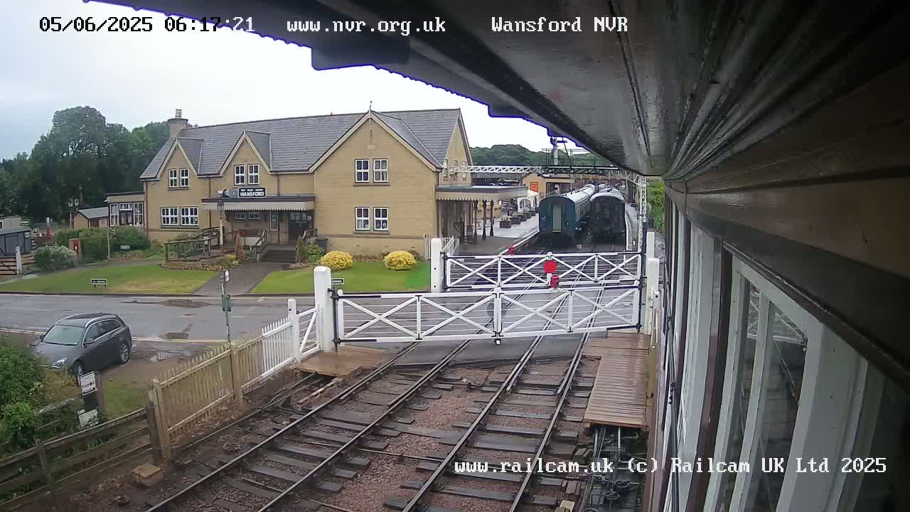A view from a train shows a railway station building and train cars under an overcast sky, with a car parked nearby and level crossing gates closed.
