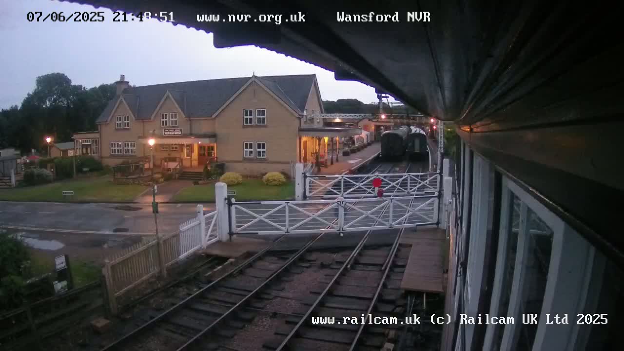A dark-colored train is at a station near a building with lights on, under an overcast sky.
