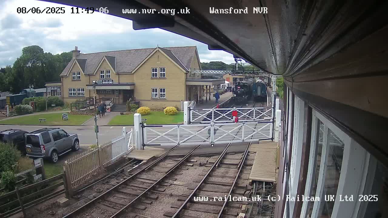A partly cloudy day at a train station with a steam train and several cars parked nearby.