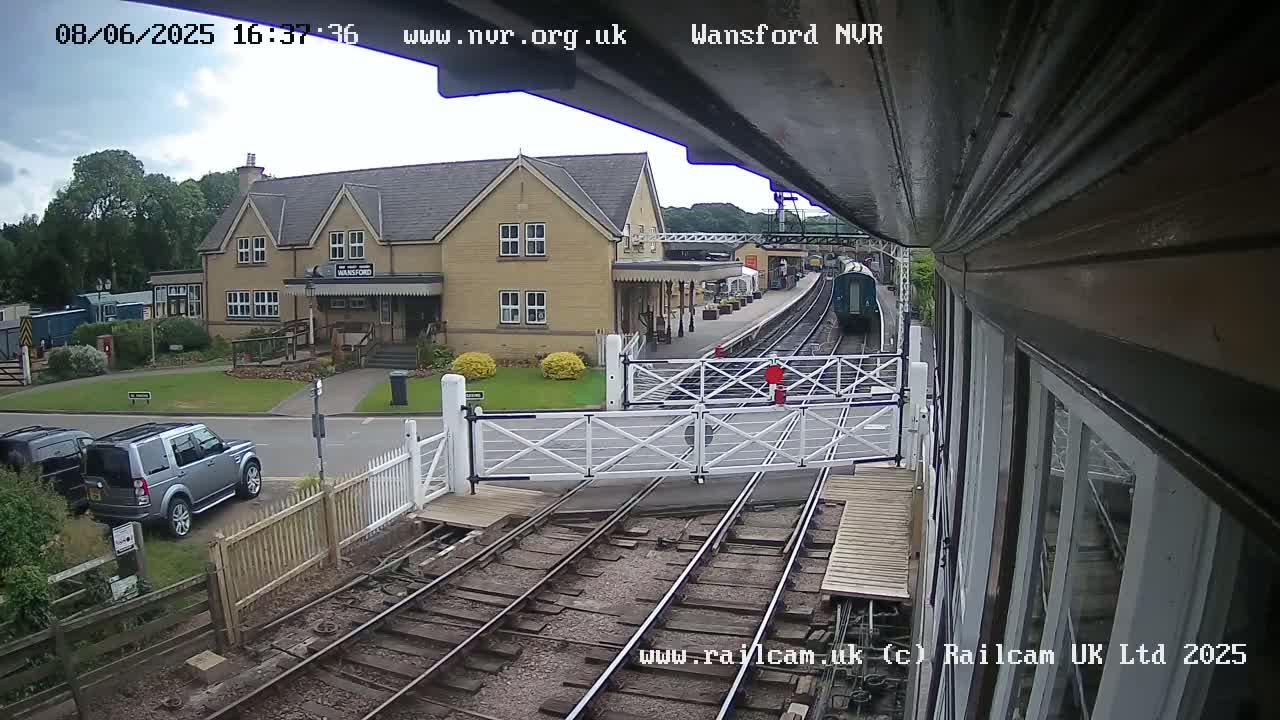 A train approaches a railway station on an overcast day, with cars parked nearby and a building visible in the background.