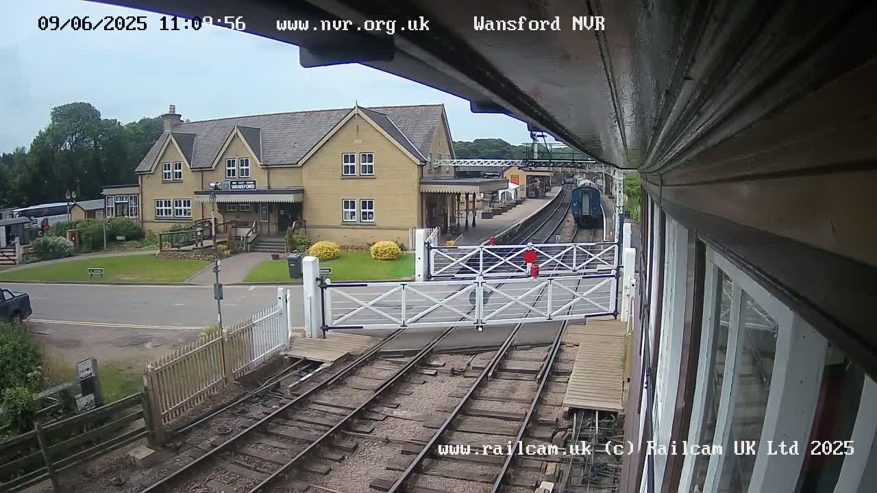 A train is approaching a station on a partly sunny day, passing through a level crossing gate in front of a light-colored building.