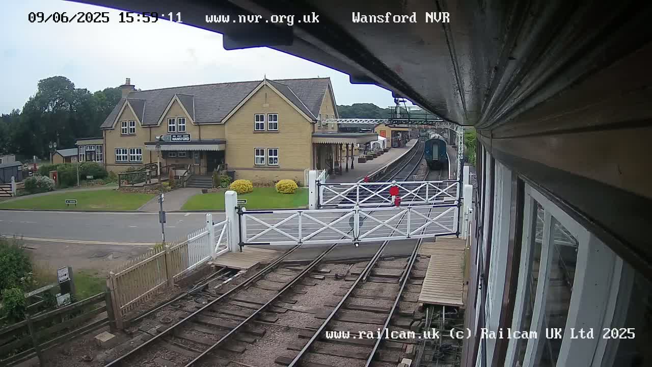 A train approaches a railway station on a partly sunny day, passing through a level crossing.