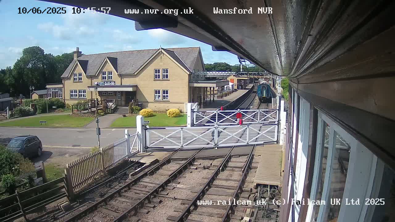 A train approaches a station on a sunny day, passing through a level crossing in front of a light-colored building.